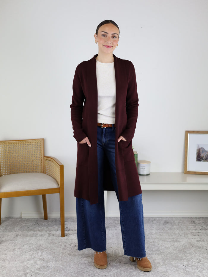 A person stands indoors wearing a cozy fall outfit, featuring the Parkland Long Cardigan in maroon over a cream top, paired with wide-leg blue jeans and brown shoes. Behind them is a woven chair and a bench displaying a candle and framed art.