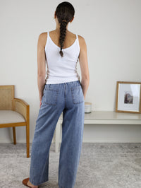Wearing the Marigold Acid Wash Wide Leg Denim and a white tank top, a person with a braid stands with their back to the camera in a minimal room featuring a chair, console table, and framed picture.