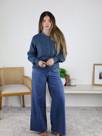 A woman with long brown hair stands indoors wearing a Theo Sweater and wide-leg jeans, holding a phone. Behind her are a chair, white bookshelf with books, a plant, and a framed photo.