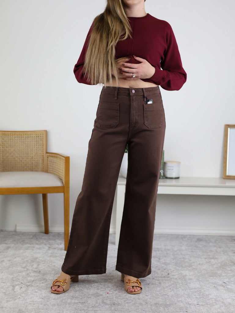A woman stands indoors wearing the Aggie Patch Pocket Wide Leg jeans, a burgundy long-sleeve top, and tan heeled sandals. Her right hand is on her waist; a chair and framed photo are in the background.