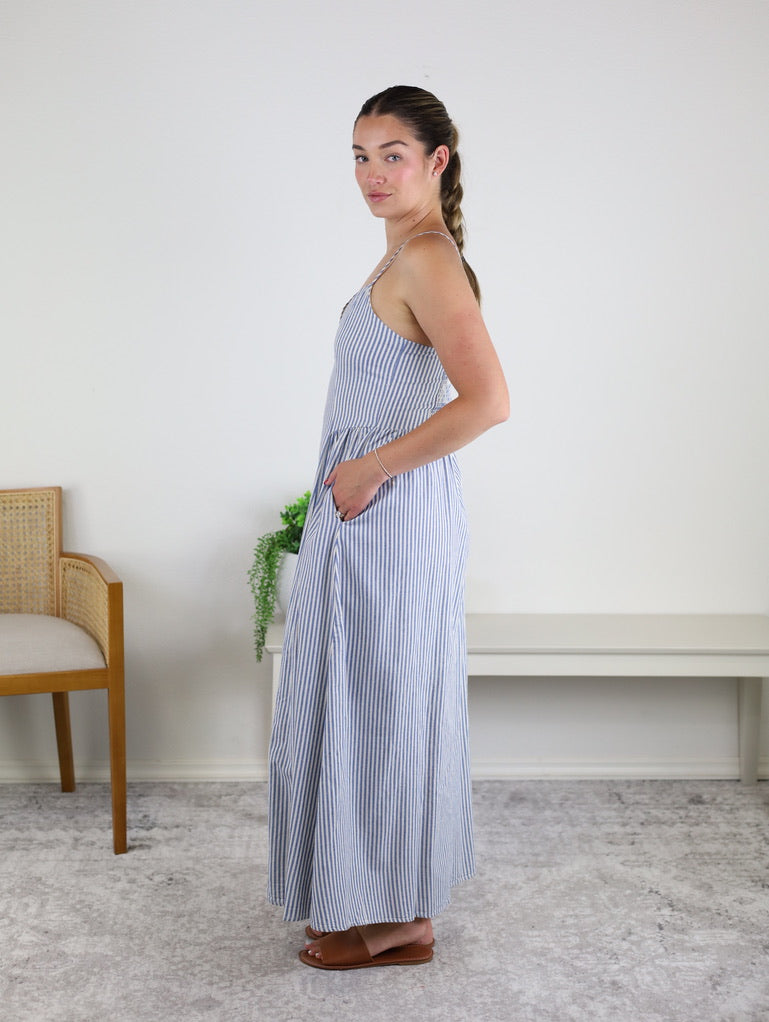 A woman stands sideways indoors by a bench and a wooden chair with a plant in the background, wearing the Molly Striped Maxi Dress—a blue and white striped maxi dress with adjustable straps and pockets—paired with brown sandals and her hair styled in a braid.