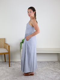 A woman stands sideways indoors by a bench and a wooden chair with a plant in the background, wearing the Molly Striped Maxi Dress—a blue and white striped maxi dress with adjustable straps and pockets—paired with brown sandals and her hair styled in a braid.