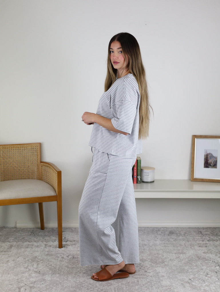 A woman with long hair wears brown sandals and the Alanna Striped Knit Top and Pants Set, standing sideways in a minimalist room with a chair, shelf, framed photo, and candle.