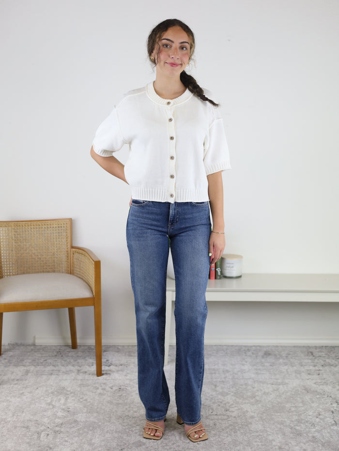A woman wearing the Nathan Raw Edge Short Sleeve Cardigan in white, paired with blue jeans and sandals, stands in a room featuring a cane chair, a white wall, and a small shelf holding candles and books. She smiles gently with one hand by her side and the other behind her back.