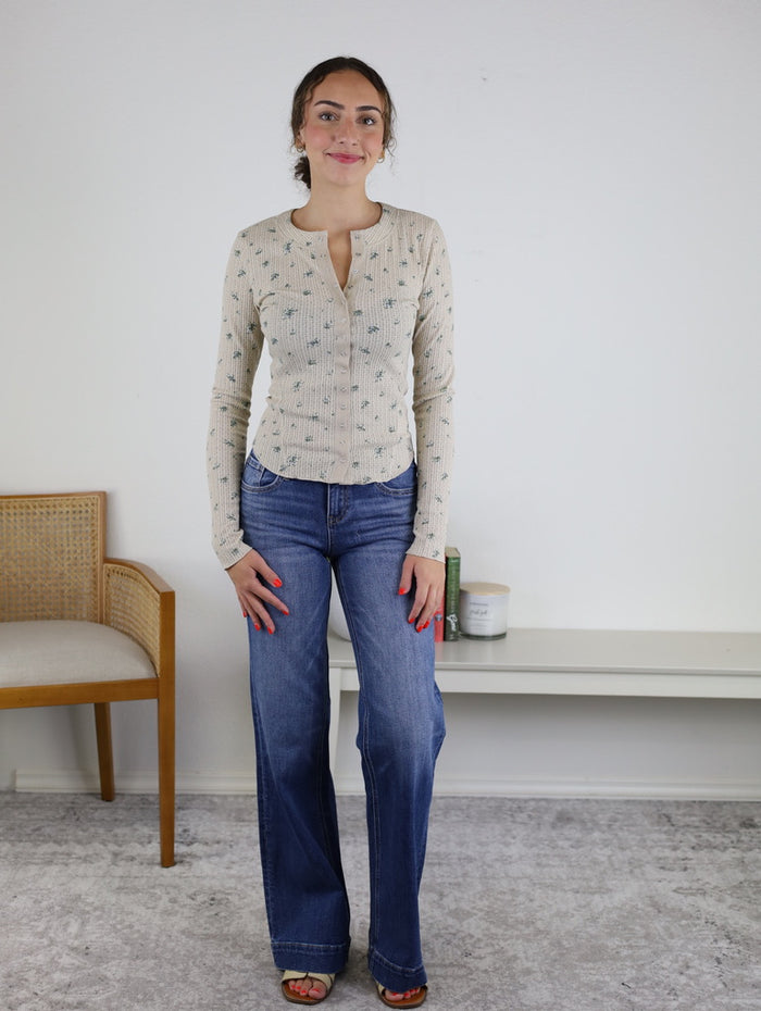 A woman stands indoors on a light carpet, wearing the Izzy Floral Waffle Knit top and blue jeans. There is a chair to her left and a white bench with books and a candle behind her.