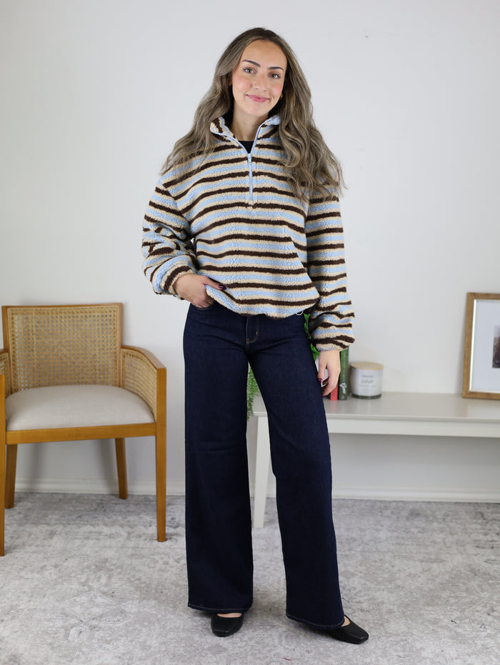 A woman wearing a tan, blue, and brown Coastal Stripe Sherpa Striped Half Zip Pullover paired with wide-leg dark wash jeans.