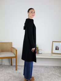 A woman stands sideways, smiling, wearing the Pinch Me Long Cardigan in black layered over blue jeans and tan shoes. She is indoors near a chair and a minimalist shelf with a plant and framed photo, perfectly embodying cozy layering for any season.