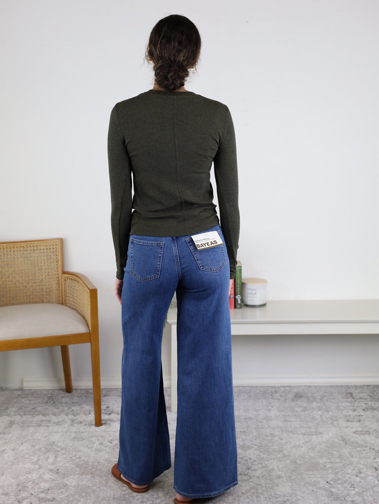 Wearing the Josette Ribbed Classic Crewneck in dark green and wide-leg blue jeans, a person stands indoors facing away. A beige chair is to the left, while a white shelf with decor items is on the right.