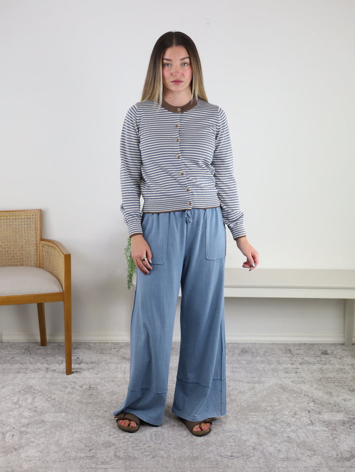 Certainly! Here’s the revised sentence using your product data: A woman stands indoors on a light gray rug, styled in the Hazel Striped Cardigan—a cozy layer crafted from lightweight polyester knit—paired with loose blue pants. In the background, a wooden chair and white bench complete the setting.