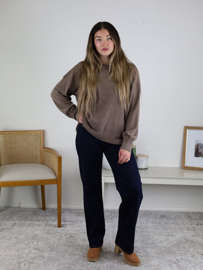 A woman with long brown hair stands indoors wearing the Lane Exposed Seam Top, dark pants, and tan heels. She poses with one hand on her hip; a chair, table, and framed picture are in the background.