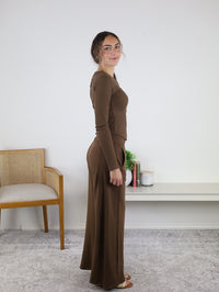 A woman stands sideways wearing the Statler Lounge Set, featuring a brown henley top and matching wide-leg pants. She smiles slightly in a simple room furnished with a chair, a book-filled shelf, a plant, and a candle.