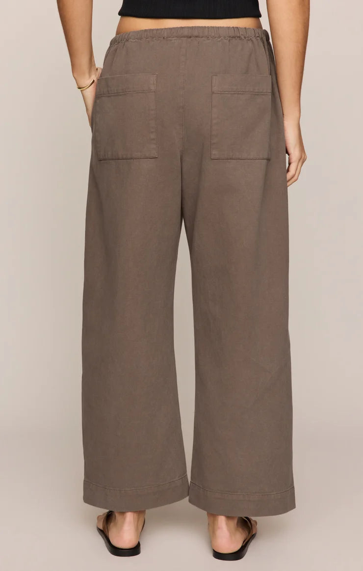 A person is shown from the back wearing the Kit Twill Carpenter Pant in Pebble—a high-waisted, wide-leg brown twill pant featuring utility pockets and two back pockets—styled with a black top and black sandals, standing against a neutral background.