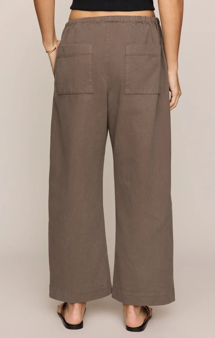 A person is shown from the back wearing the Kit Twill Carpenter Pant in Pebble—a high-waisted, wide-leg brown twill pant featuring utility pockets and two back pockets—styled with a black top and black sandals, standing against a neutral background.