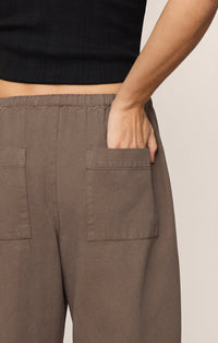 A person wearing the Kit Twill Carpenter Pant in Pebble and a black top places their right hand in a back utility pocket. The image is cropped to show only the lower back and part of the arm.
