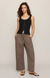A woman stands against a plain background wearing the Kit Twill Carpenter Pant in Pebble, styled with a sleeveless black button-down top and black sandals, with one hand in her pocket.