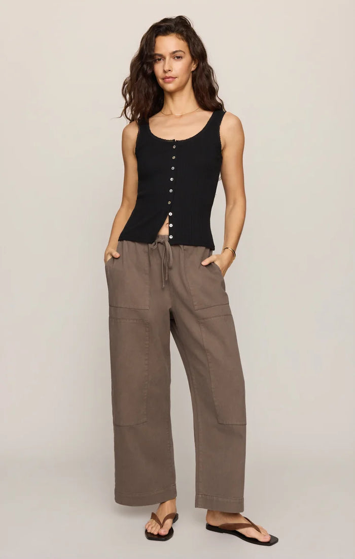 A woman stands against a plain background wearing the Kit Twill Carpenter Pant in Pebble, styled with a sleeveless black button-down top and black sandals, with one hand in her pocket.