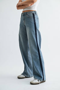 A person is wearing the JBD - Mid Rise Sleek Tuxedo Sweat Pants in Medium Denim (DP914: 24), featuring a light blue wash and a dark stripe down the side for a tuxedo-inspired style. The pants are paired with a white cropped top and sneakers, and the person stands with arms crossed against a plain white background.