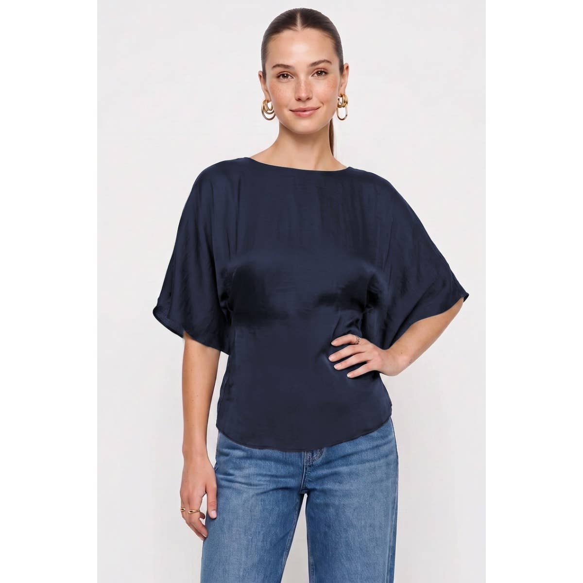 A woman wearing a Naked Zebra - Back Tie Dolman Sleeve Silky Fall Blouse: Wine / L with a dark navy blue silky dolman sleeve blouse with a boat neckline and blue jeans.