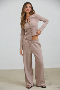 A woman wearing the Creamon - CTP3592 BROWN Lounge Striped Button Down Top & Pants Set in brown stripe, featuring a button-down top and drawstring wide leg pants, stands on a light-colored floor against a plain white wall.