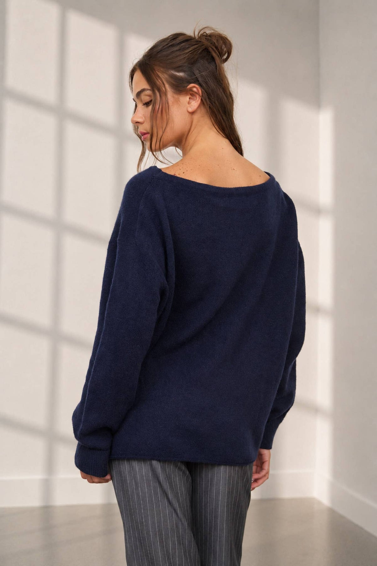 A woman with brown hair in a messy bun, wearing The Workshop Studio’s Boat Neck Oversized Knit Pullover Sweater in Red Wood and gray pinstripe pants, stands in a brightly lit room facing away as sunlight streams through large window panes.
