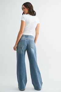 A woman stands with her back to the camera against a plain light background, wearing a white fitted t-shirt paired with JBD - Mid Rise Sleek Tuxedo Sweat Pants in medium denim.