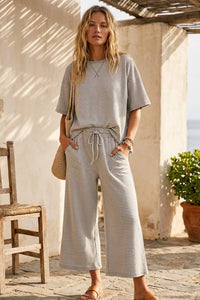 Outdoors, a woman wears the Alanna Striped Knit Top and Pants Set—loose short-sleeve top and wide-leg pants—with sandals and a woven bag, standing by a wooden chair against a sunlit wall, hands in her pockets.