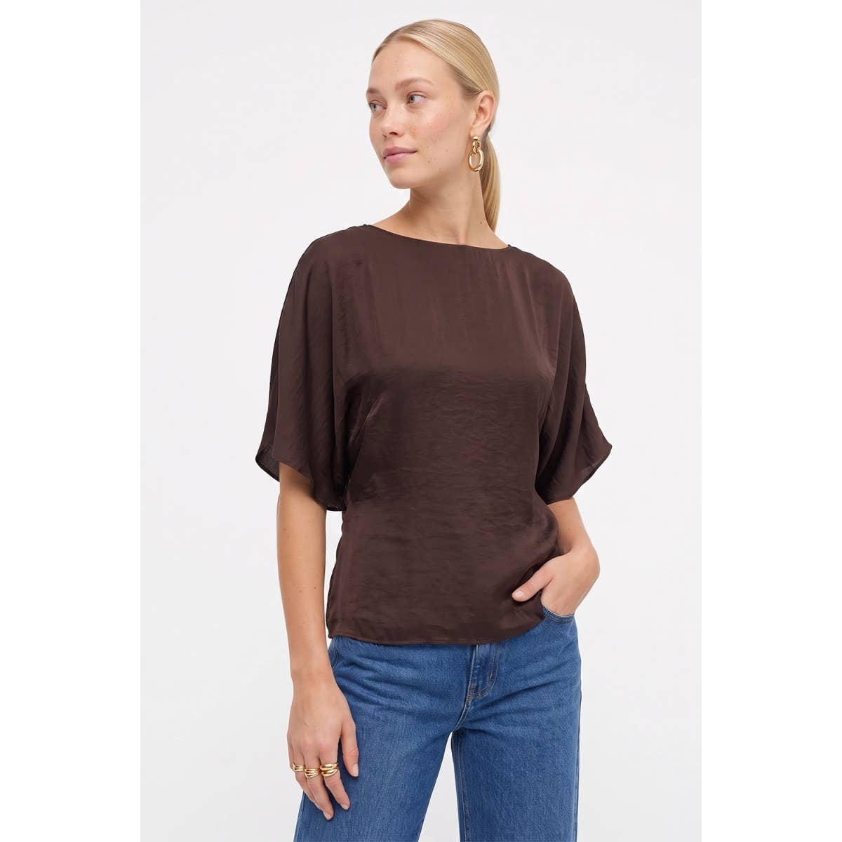 A woman wears a Naked Zebra - Back Tie Dolman Sleeve Silky Fall Blouse in dark navy with a boat neckline and blue jeans.