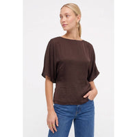 A woman wears a Naked Zebra - Back Tie Dolman Sleeve Silky Fall Blouse in dark navy with a boat neckline and blue jeans.