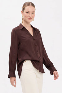 A woman wears a Naked Zebra - Neck Shirt With Open Seam Sleeve: Black / L in dark brown woven polyester with open seam sleeves, paired with white jeans.