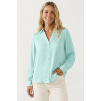 A smiling woman wears a light mint green, long-sleeved Naked Zebra - Neck Shirt With Open Seam Sleeve: Black / L tucked into white pants.