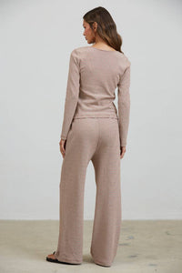 A woman with long brown hair stands indoors, facing away, wearing the Creamon - CTP3592 BROWN Lounge Striped Button Down Top & Pants Set in Brown Stripe. The set features a long-sleeve button down top and matching wide leg pants with subtle stripes. She is standing on a light-colored floor against a plain white wall.