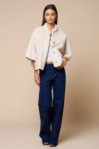 A person wears a cream-colored Jordan Fur Cape Sleeve Jacket over a white top and dark blue wide-leg jeans.
