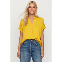 A woman wearing a Naked Zebra - Drop Shoulder Regular Blouse: Chocolate / L, a mustard yellow, short-sleeved, drop-shoulder blouse with a v-neckline, and blue jeans.