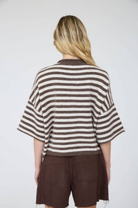 A person with long blonde hair, seen from behind, wears the Stephanie Striped Cardigan—a boxy brown and white striped wool-blend knit—paired with dark brown shorts against a plain light background.