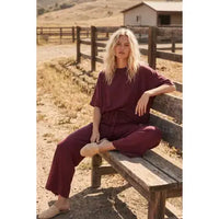 A woman with wavy blonde hair sits on a wooden bench outdoors, wearing the Heather Brushed Stretch Boxy Top & Wide Leg Pant Set—an effortlessly comfortable loungewear ensemble made from brushed stretch fabric. Behind her, dry grass, wooden fences, and a barn are visible beneath a clear sky.
