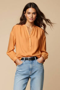 A woman wearing a Naked Zebra - Button-down Blouse With Gathered Neckline in orange, styled with long sleeves, tucked into blue jeans with a black belt.