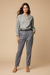 A woman wears an elegant, grey, long-sleeved Naked Zebra - Button-down Blouse With Gathered Neckline with a gathered neckline tucked into grey pleated trousers with a black belt.
