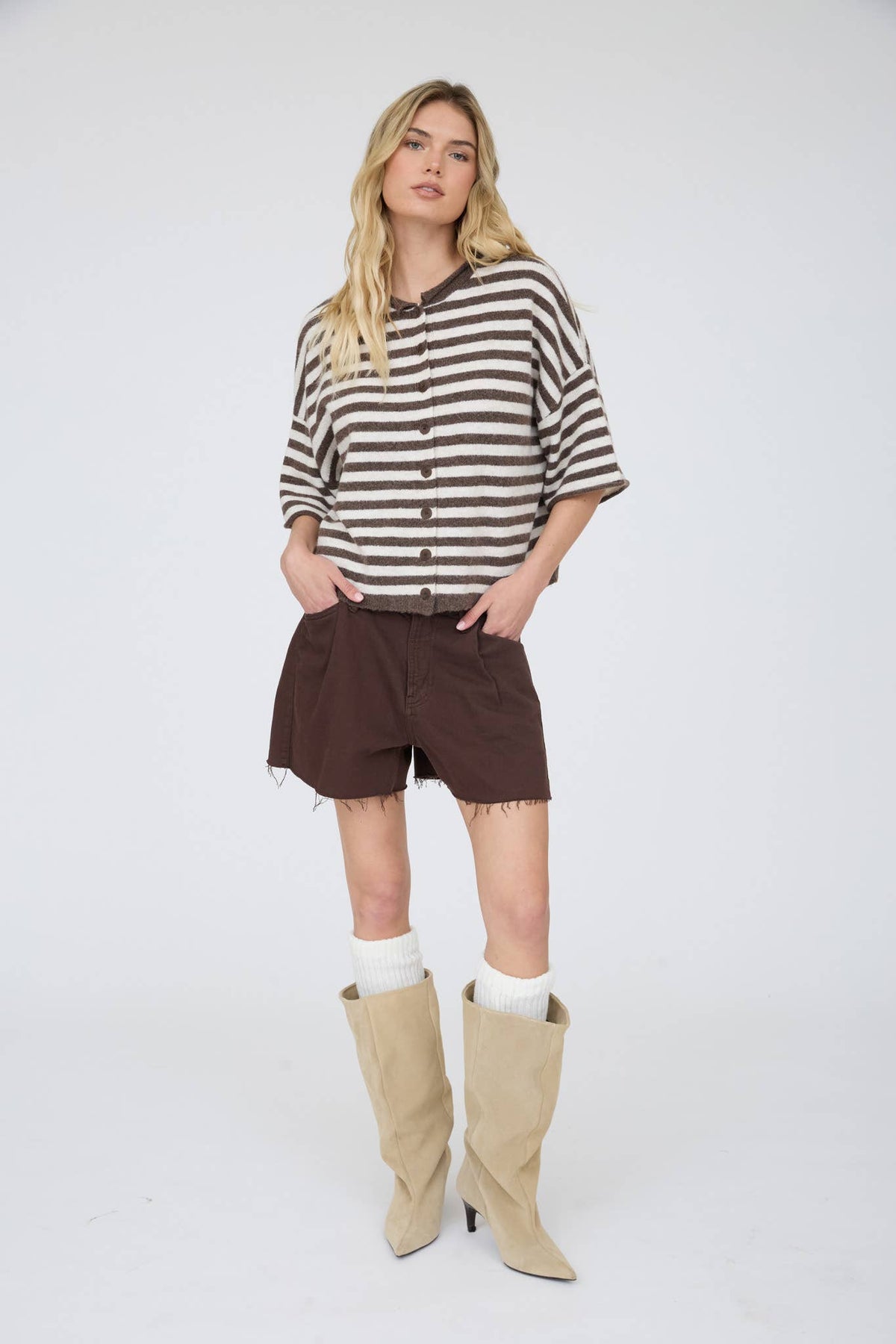 A woman with long blonde hair stands against a white background, wearing the Stephanie Striped Cardigan in chocolate brown, paired with dark brown shorts, white socks, and beige knee-high boots. Her hands are tucked in her shorts pockets.