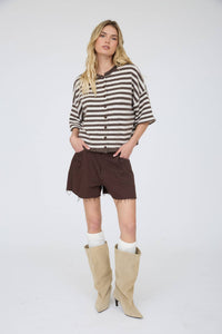 A woman with long blonde hair stands against a white background, wearing the Stephanie Striped Cardigan in chocolate brown, paired with dark brown shorts, white socks, and beige knee-high boots. Her hands are tucked in her shorts pockets.