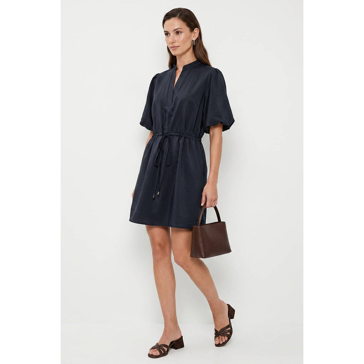 A woman wears a Naked Zebra - Puff Sleeve Cinched Waist Mini Dress: Navy / L, paired with brown heeled sandals and a matching brown handbag.