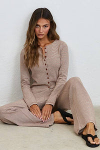 A woman with long wavy hair sits on the floor against a white wall, wearing the Creamon - CTP3592 BROWN Lounge Striped Button Down Top & Pants Set in brown stripe, paired with black sandals.