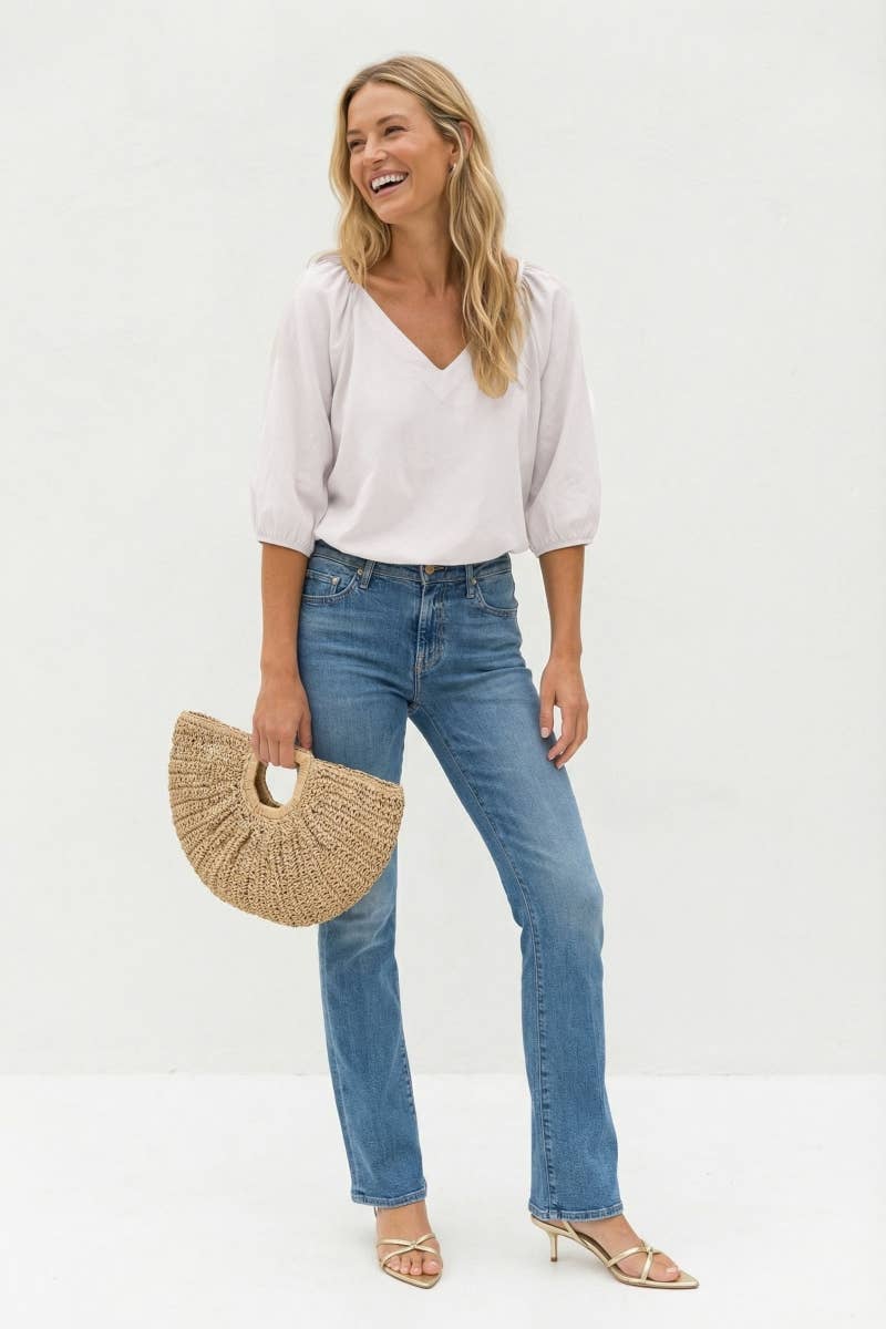 A woman wearing a Naked Zebra - Elastic Puff Sleeve V-neck Blouse in burgundy, blue jeans, and gold strappy heels, holding a crescent-shaped straw handbag.
