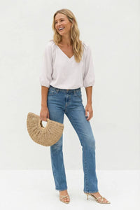 A woman wearing a Naked Zebra - Elastic Puff Sleeve V-neck Blouse in burgundy, blue jeans, and gold strappy heels, holding a crescent-shaped straw handbag.