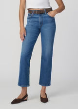 Model wears The Sienna Straight jeans in a vintage-inspired wash, styled with a brown belt, white top, and brown pointed flats, standing with one hand in the pocket against a plain light background.