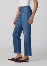 A person wearing The Sienna Straight jeans in a vintage-inspired wash, paired with a white sleeveless top and black flat shoes, stands against a plain light background.
