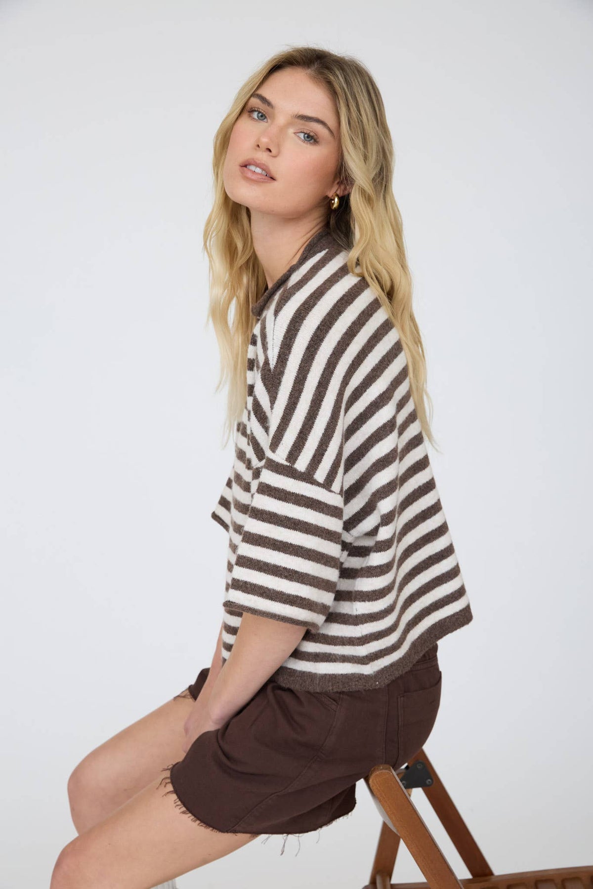A woman with long blonde hair sits sideways on a wooden stool, wearing the Stephanie Striped Cardigan and brown shorts, looking at the camera with a neutral expression against a plain background.