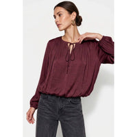 A woman wears a Naked Zebra - Long Sleeve Draped Bubble Hem Blouse: Wine / L featuring a tie-neck detail and gathered bubble hem, paired with dark grey jeans.