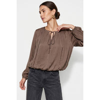 A wine Naked Zebra - Long Sleeve Draped Bubble Hem Blouse featuring a gathered neckline, tie detail, and bubble hem, worn with dark gray jeans.