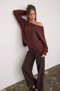 A woman poses against a white wall wearing The Workshop Studio’s Boat Neck Oversized Knit Pullover Sweater in Red Wood, styled in an oversized fit with brown pinstriped wide-leg pants, one arm raised and her gaze directed downward.
