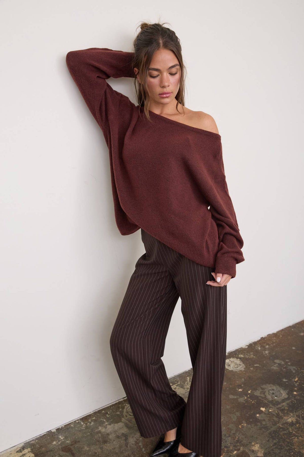 A woman poses against a white wall, wearing The Workshop Studio’s boat neck oversized knit pullover sweater in oatmeal and brown pinstripe pants. One arm is bent behind her head, and she looks down with a relaxed expression.